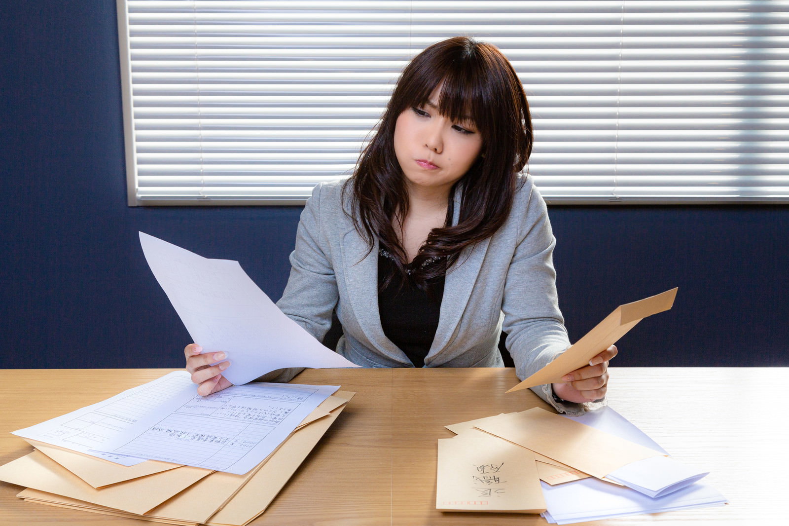 デスクで書類を手に困った表情を見せるグレーのスーツを着た女性［モデル：Lala］