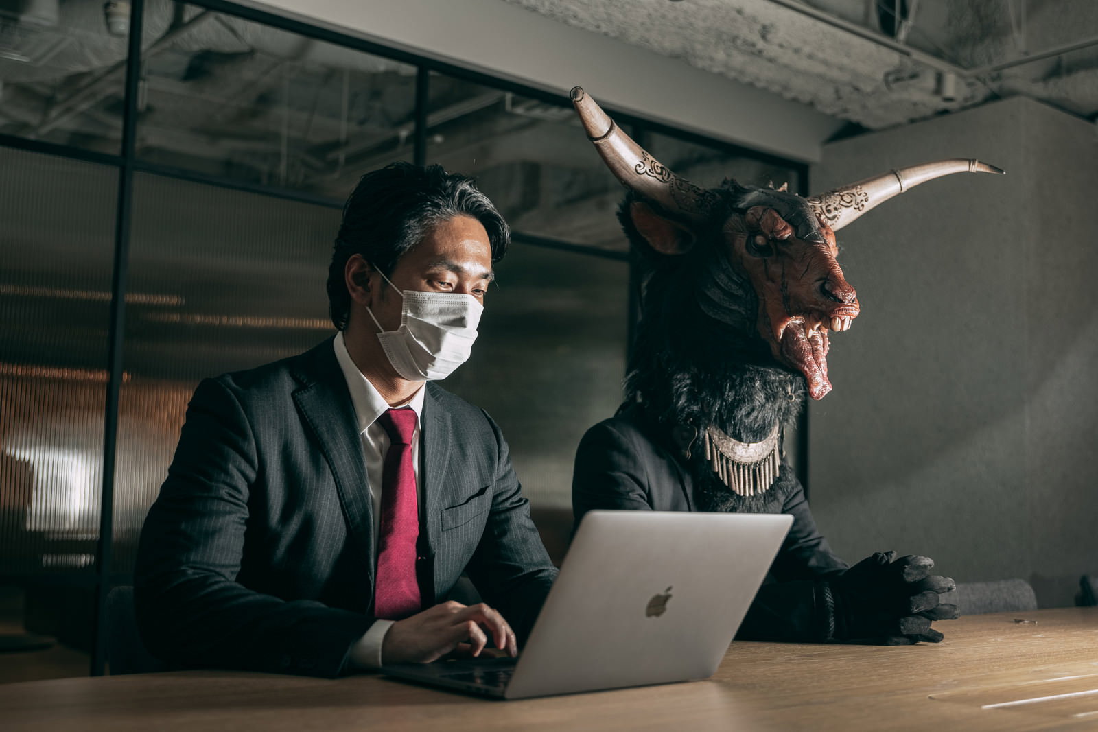 オフィスでマスクをした会社員と角の生えた魔神がパソコンで作業
