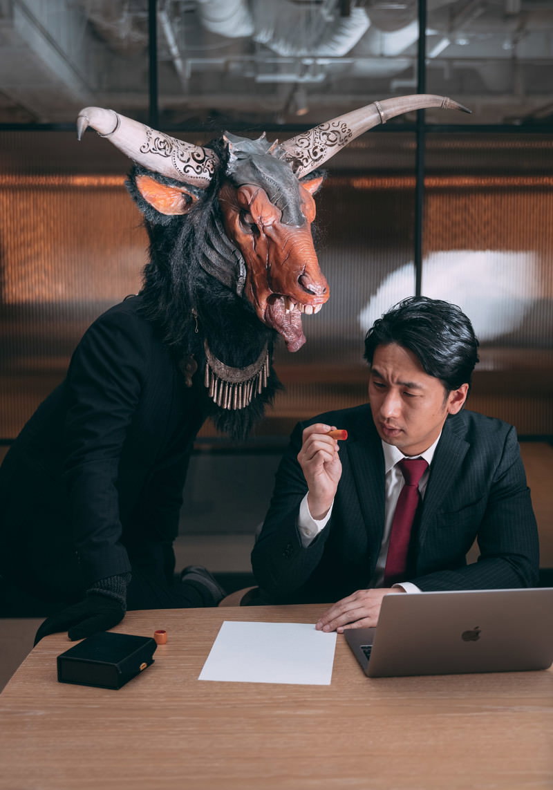 悪魔の面をつけたスーツ姿の男性がオフィスで書類を見つめている様子［モデル：大川竜弥］