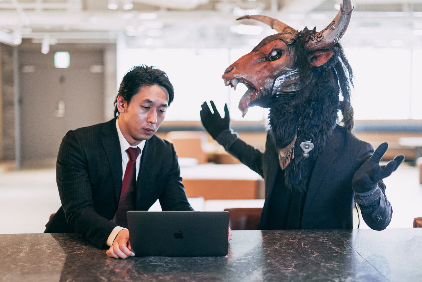 オフィスの机で、スーツ姿の男性と魔神マスク姿の上司が対面し、魔神が威圧的にジェスチャーしている