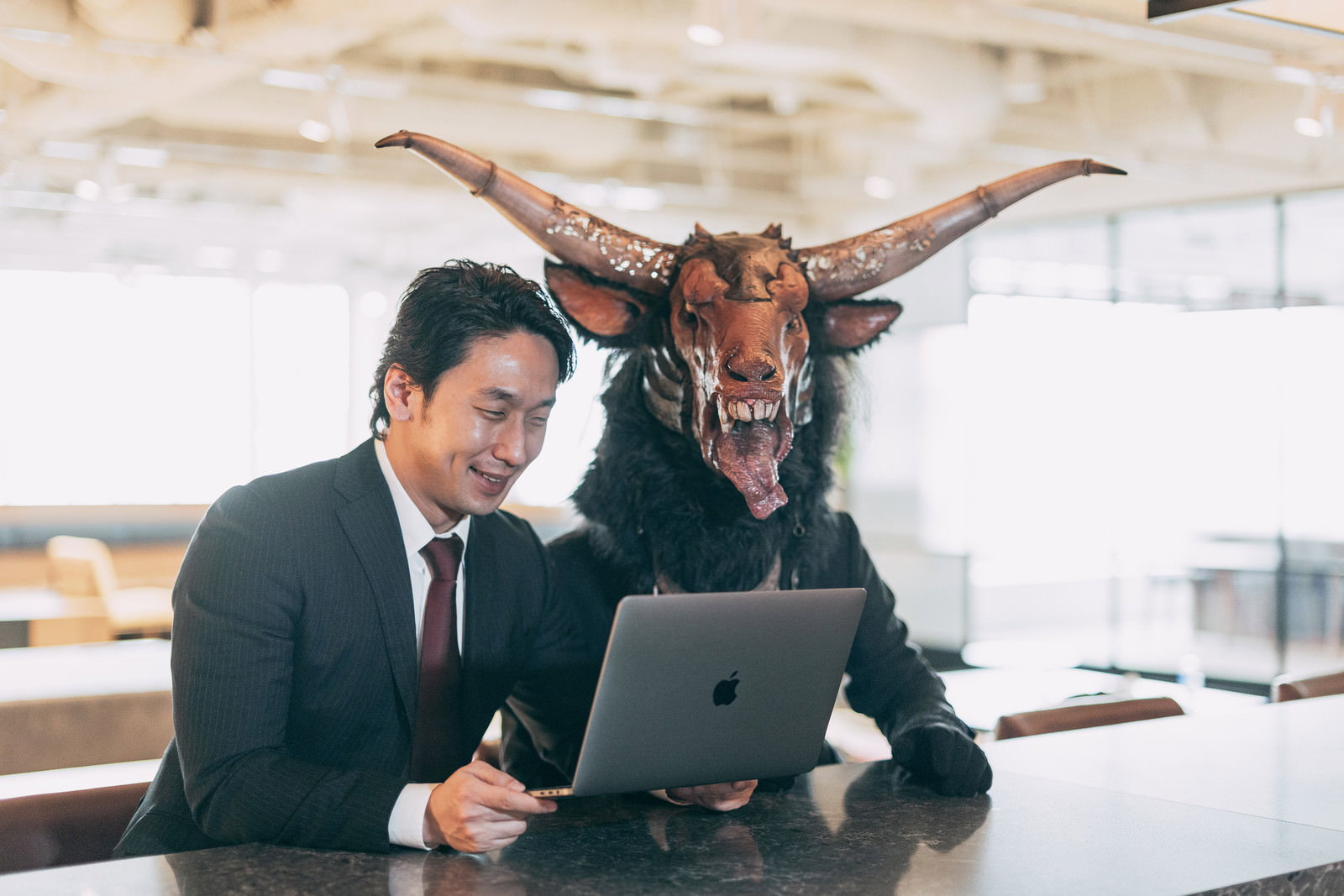 ノートパソコンを見つめる笑顔のスーツ姿男性と背後に控える角の生えた魔神