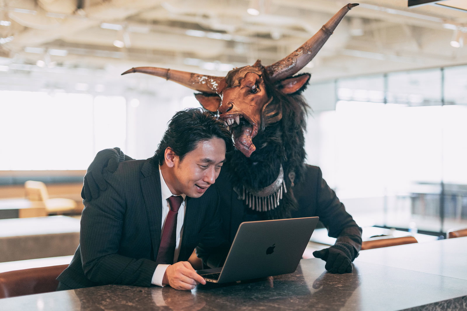 角の生えた魔神がスーツを着て、オフィスでノートパソコンを見る部下の肩に手を置いて褒めている様子
