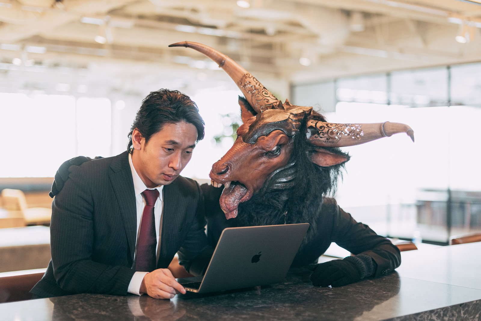 スーツ姿の男性と魔神風キャラクターが机で向かい合う様子［モデル：大川竜弥］