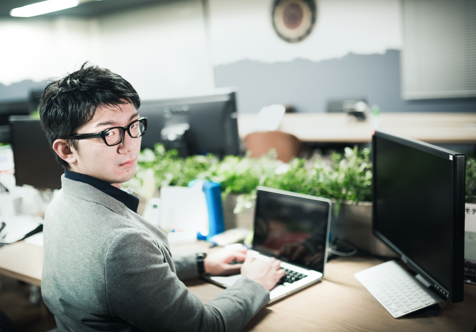 オフィスのデスクでキーボード操作しながら後ろを振り返る、黒縁眼鏡をかけたグレーシャツ姿の男性エンジニア［モデル：OZPA］