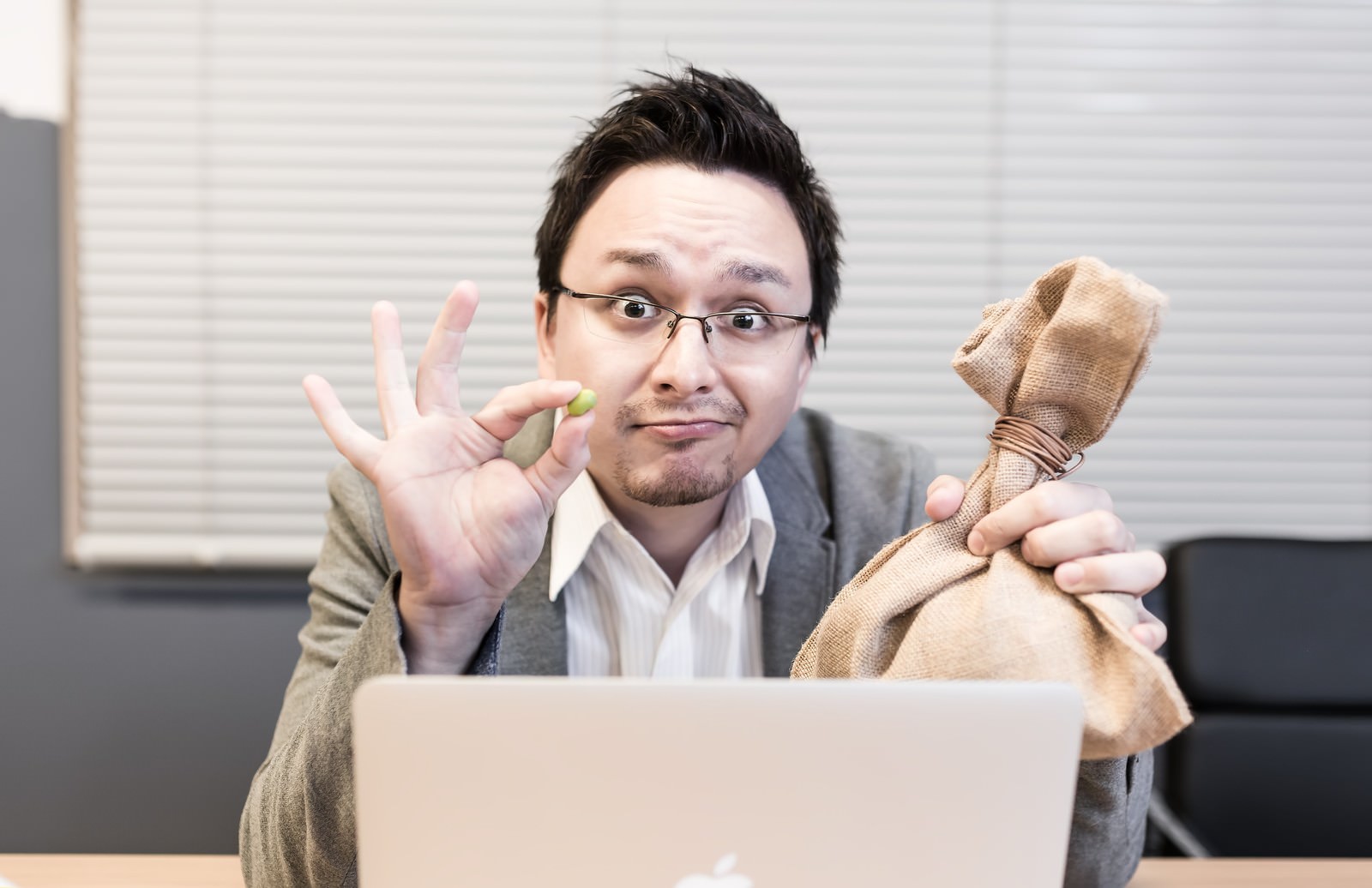 眼鏡の男性がオフィスで黄色い豆を持ち、困惑した表情で片手を上げている［モデル：Max_Ezaki］