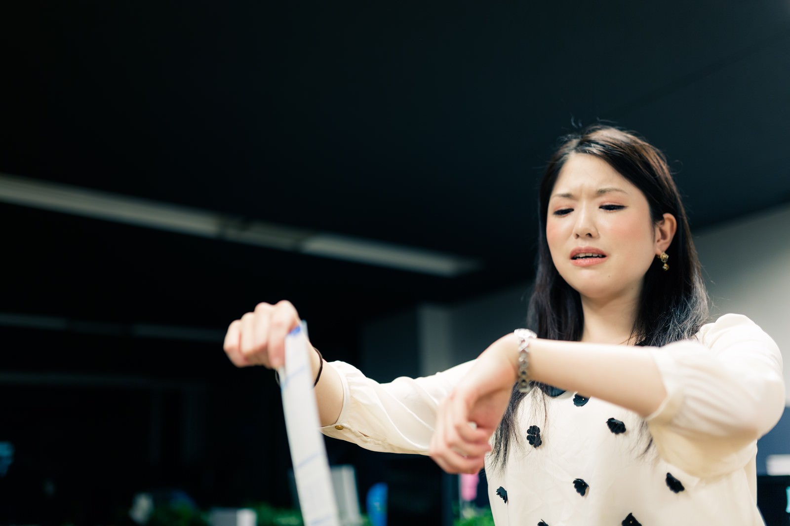 オフィスで白いドット柄ブラウスを着た女性が紙を持ち両腕を広げ、困惑した表情を浮かべている［モデル：シマヅ］