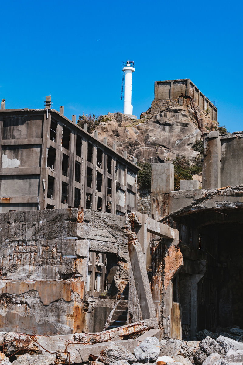 青空の下、軍艦島の高台に立つ白い肥前端島灯台と周囲の廃墟建物群