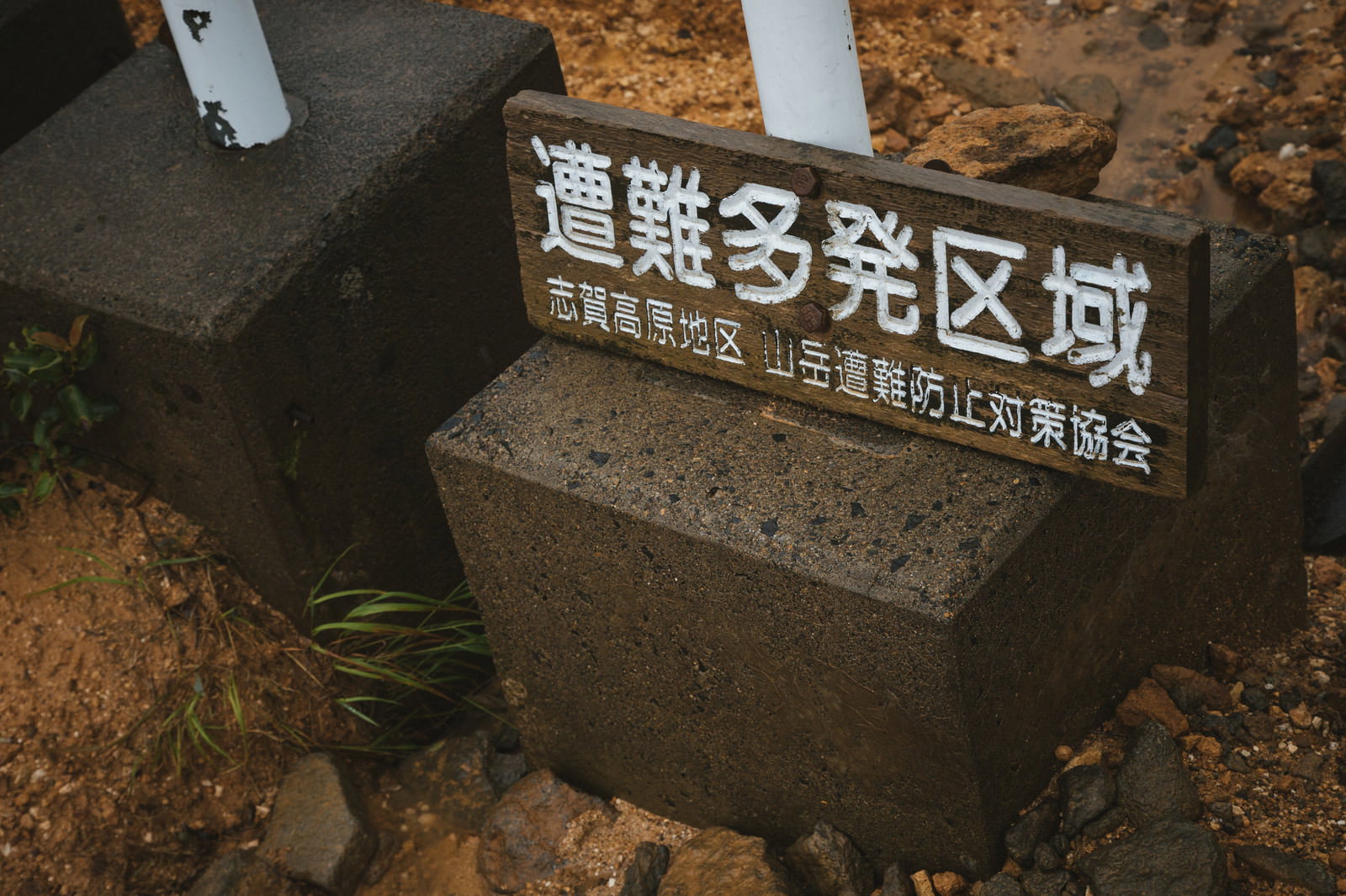 茶色い木製の警告看板に白い日本語テキストが記載され、周囲に落ち葉と土が散在している
