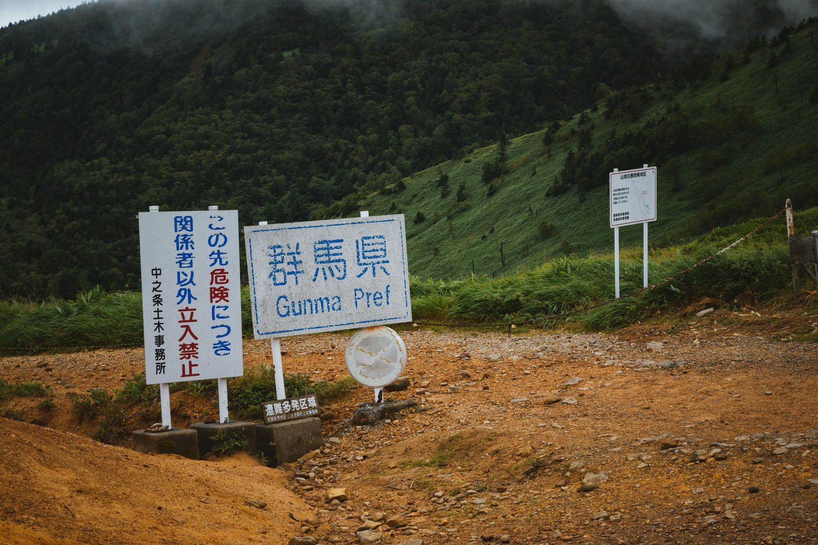 緑の山々を背景に、未舗装路脇に並ぶ複数の立入禁止看板。警告表記と指定区域を示す白い看板