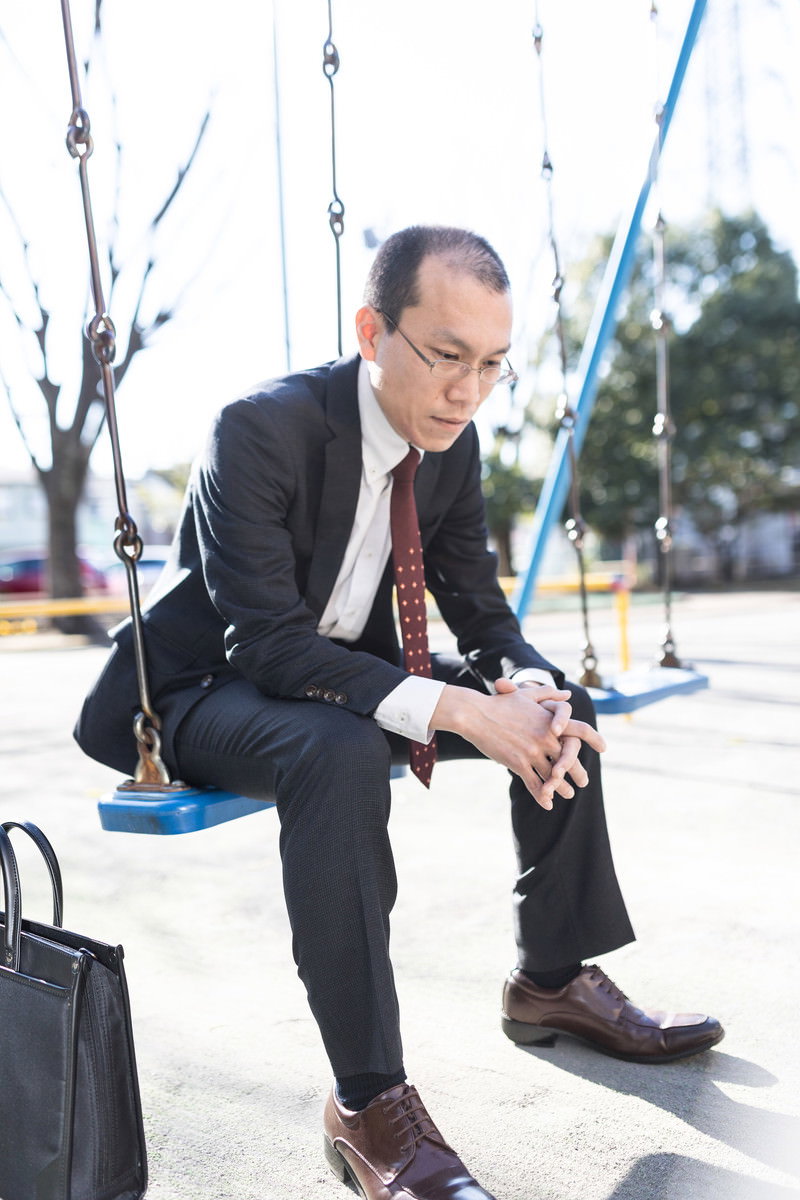 公園のブランコに座り項垂れているスーツ姿の男性［モデル：サンライズ鈴木］