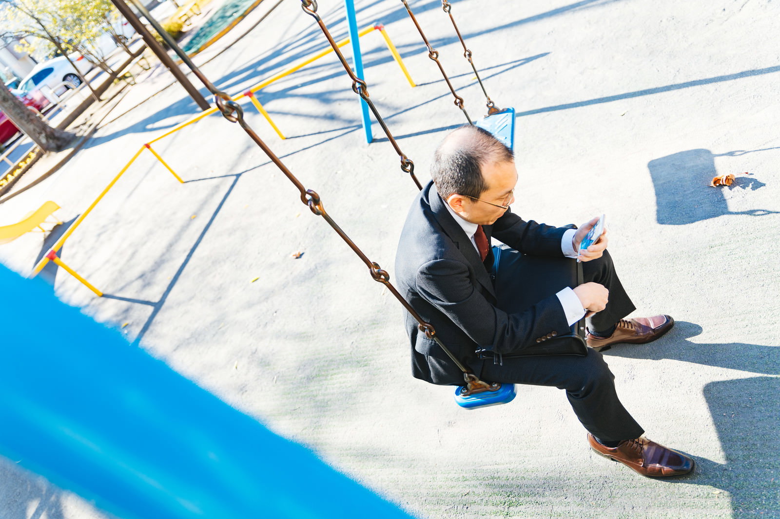 ブランコに座りスマートフォンを操作するスーツ姿の中年男性。背景に遊具が見える公園での撮影［モデル：サンライズ鈴木］