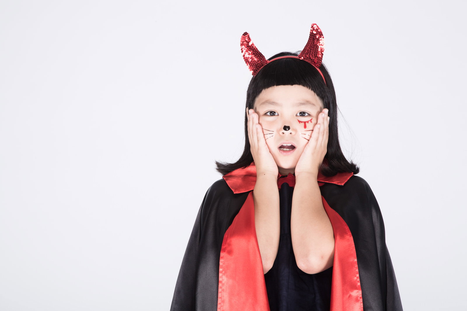 赤い角のカチューシャと黒いマントを着た女の子が頬に手を当てて驚いている様子［モデル：ゆうき］