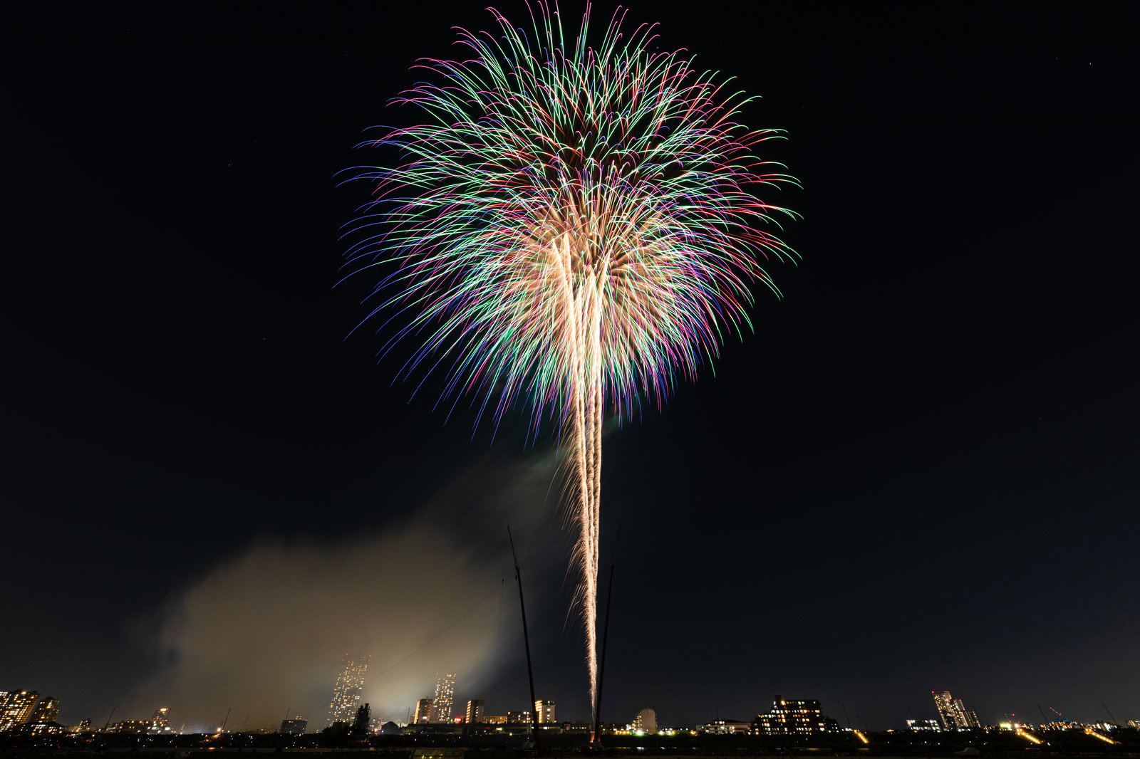 夜空に大きく開いた色とりどりの打ち上げ花火、風で光跡が流れている