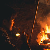 焚き火の炎の中、串焼きの焼き鳥を食らうソロキャン中の男性の写真