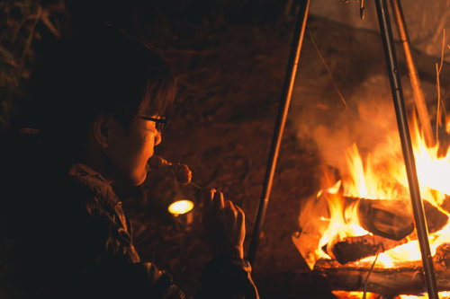焚き火の炎の中、串焼きの焼き鳥を食らうソロキャン中の男性