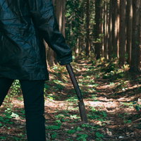 森の木立で黒い鎌を持つ作業着姿の男性の写真