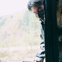 山道の古い建物の陰から周囲を伺う長髪の男性の警戒の写真