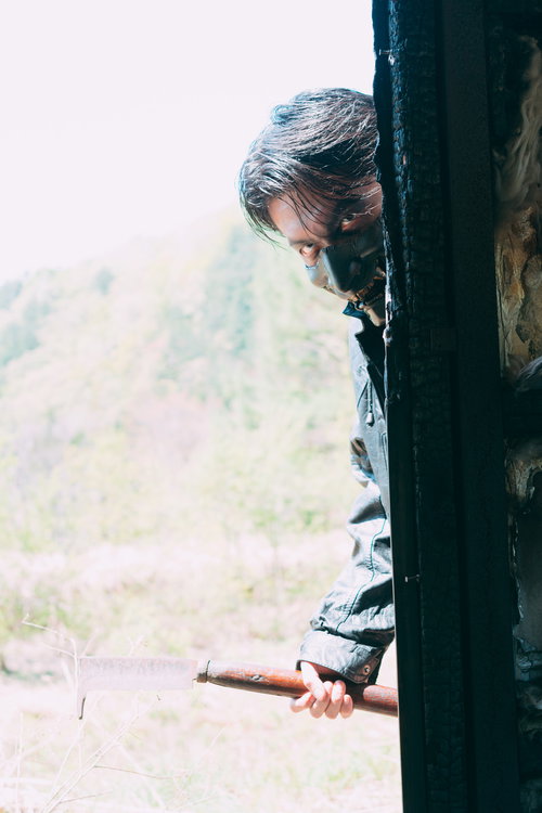 山道の古い建物の陰から周囲を伺う長髪の男性の警戒