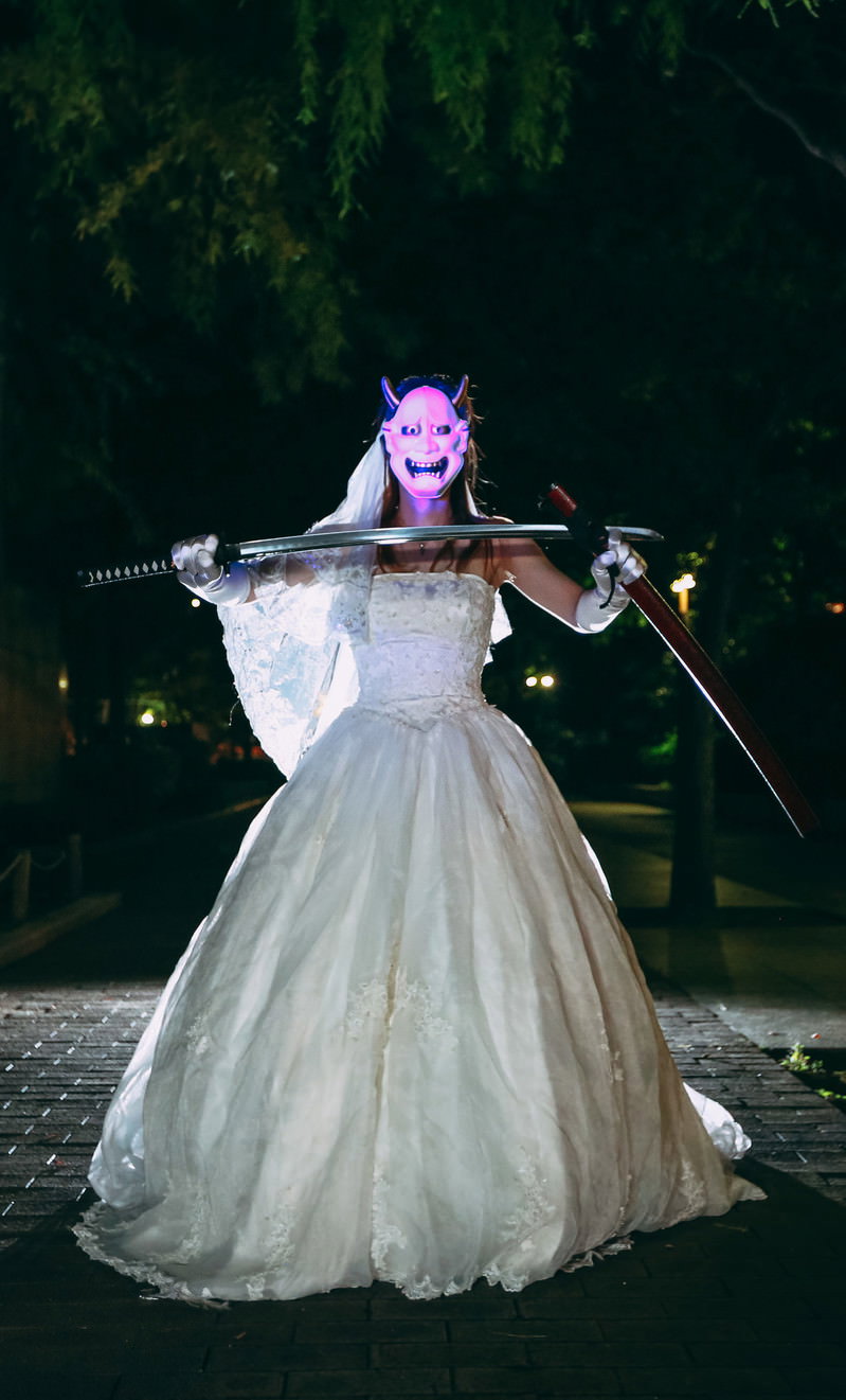 白いウェディングドレスを着て般若の面をつけた花嫁が夜道で刀を抜いている様子［モデル：モデルリリース］