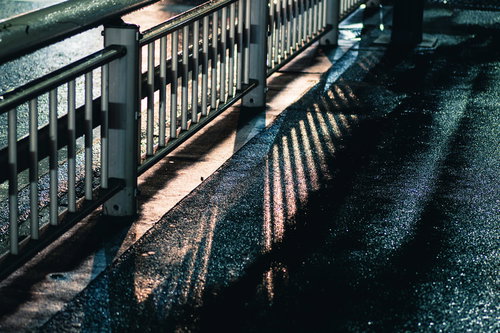 雨上がりの濡れた歩道に伸びる防護柵の影と水たまりの反射