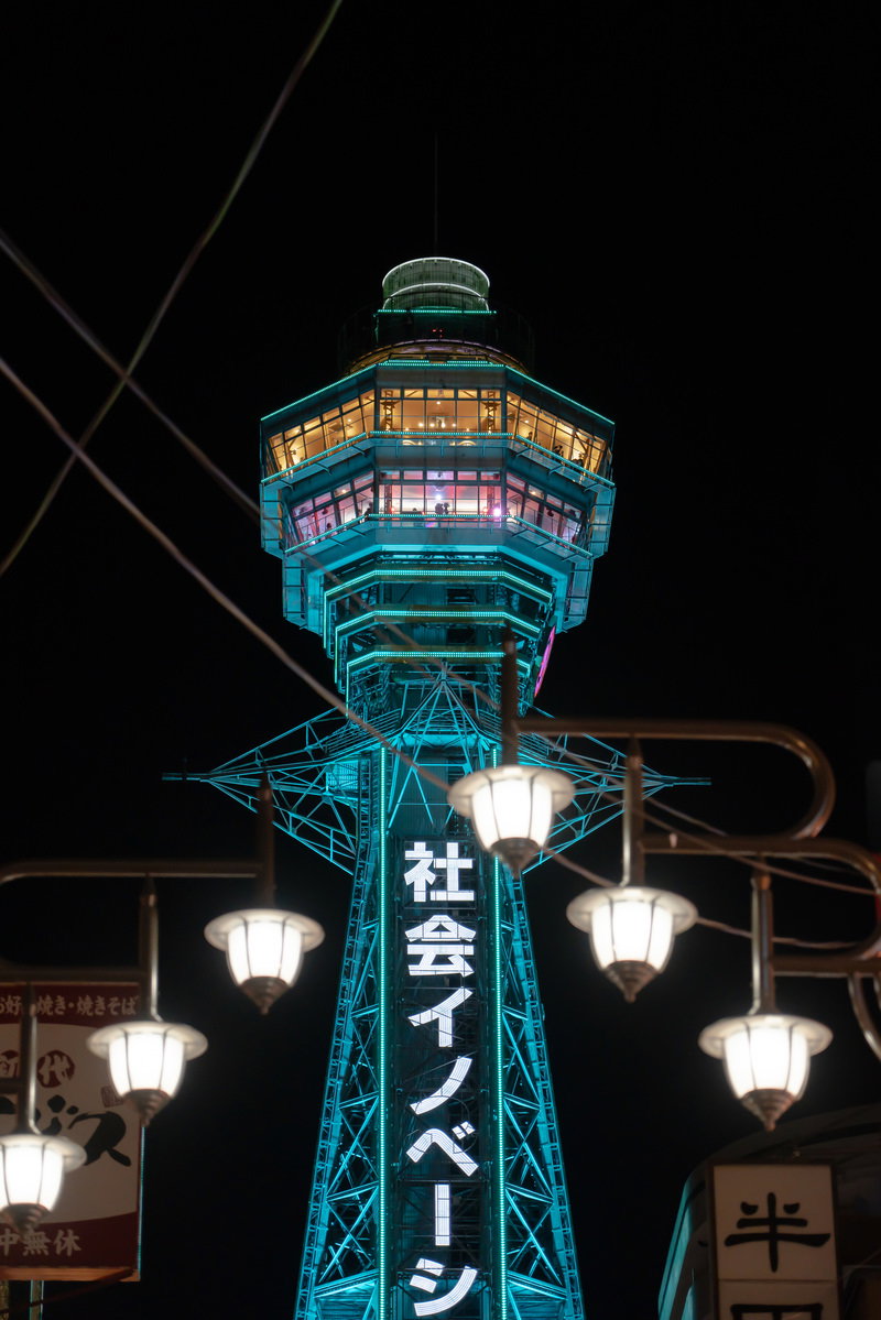 水色にライトアップされた夜の通天閣を街灯越しに見上げた写真