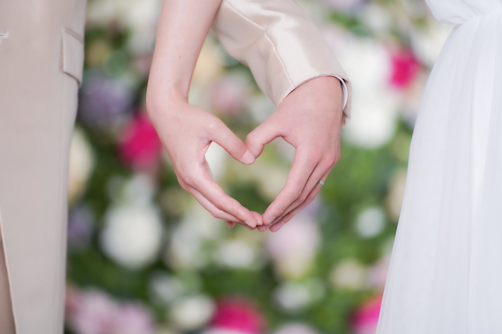 白いウェディングドレスとスーツを着た新郎新婦が手でハート形を作っている様子［モデル：たけべともこ］