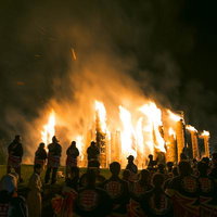 燃え上がる松明あかしを取り囲む観客たち、夜空に立ち上る炎と白煙の写真