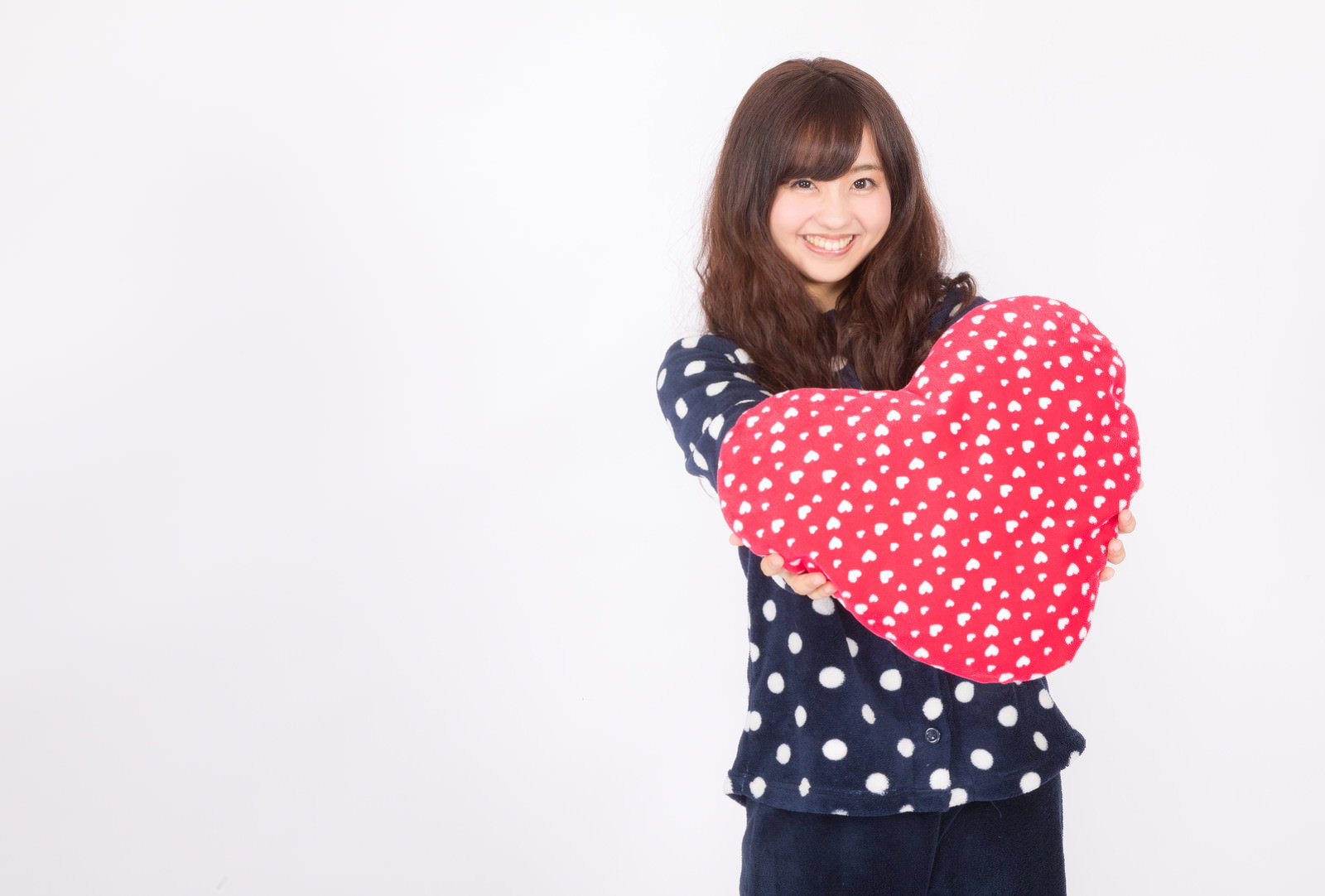 ネイビーのドット柄パジャマを着た女性が赤いハート型クッションを差し出している［モデル：河村友歌］