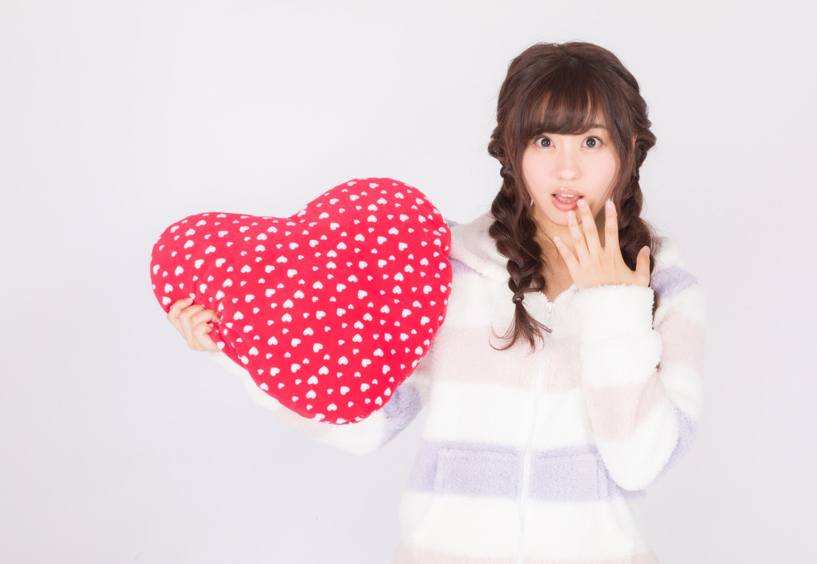 白いパジャマを着た女性がドット柄のハート型クッションを抱えて、口元に手を当てて驚いた表情をしている［モデル：河村友歌］