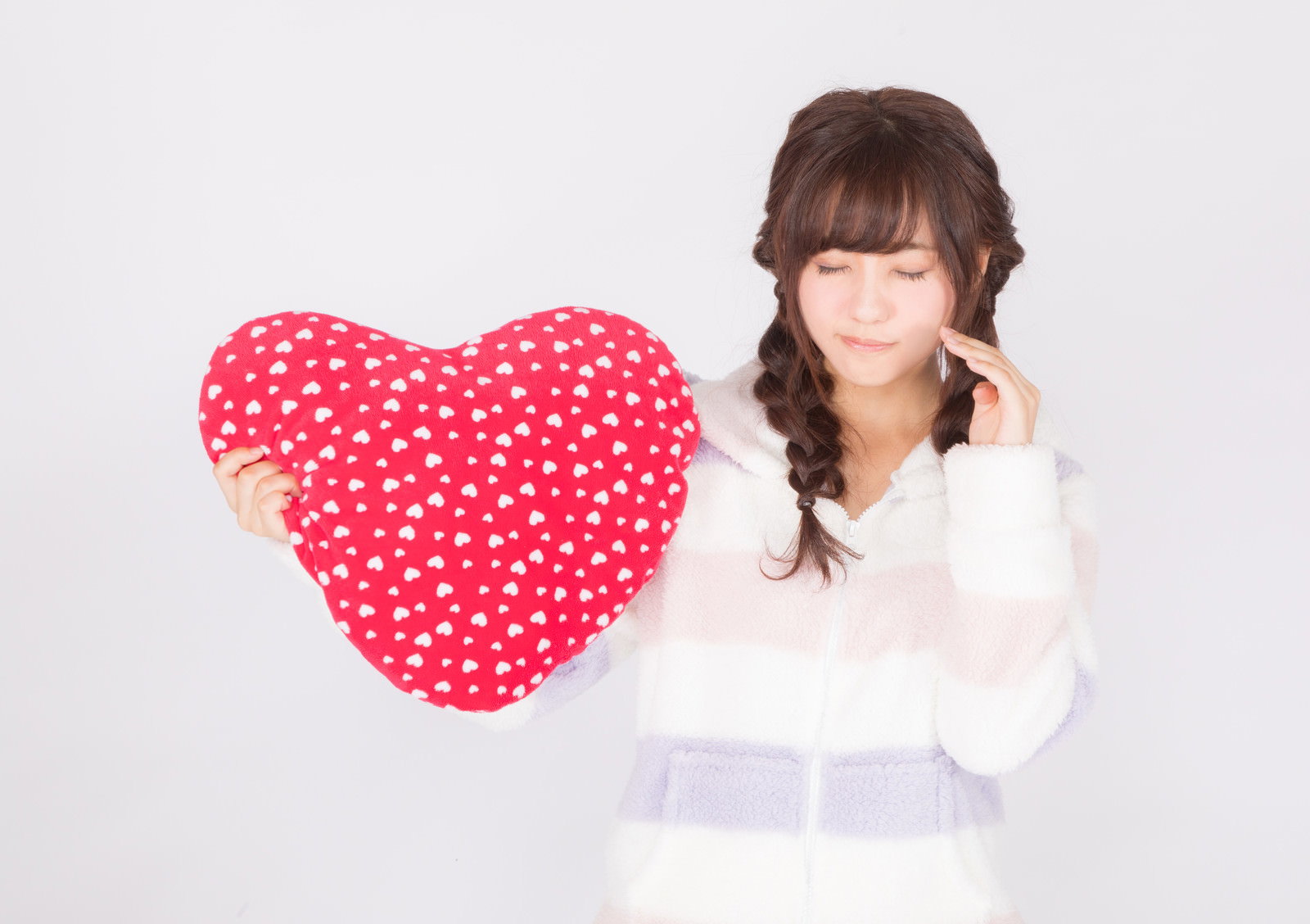 赤いドット柄のハートのクッションを両手で持つ白いパジャマ姿の女性［モデル：河村友歌］