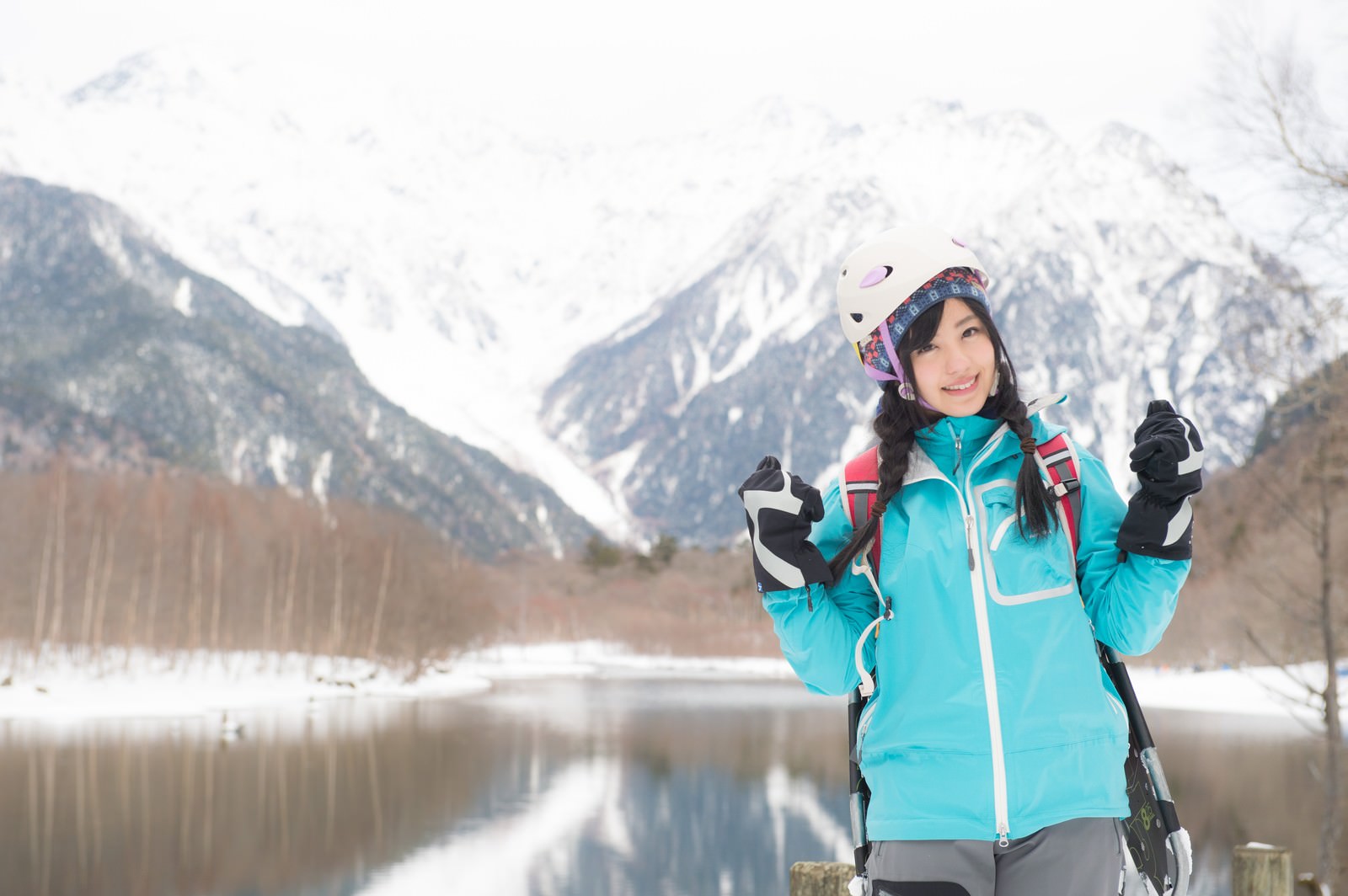 青いジャケットとヘルメット、赤いバックパックを装備した登山者が雪の上高地でガッツポーズをしており、背後に北アルプスの雪景色が広がっている［モデル：河村友歌］