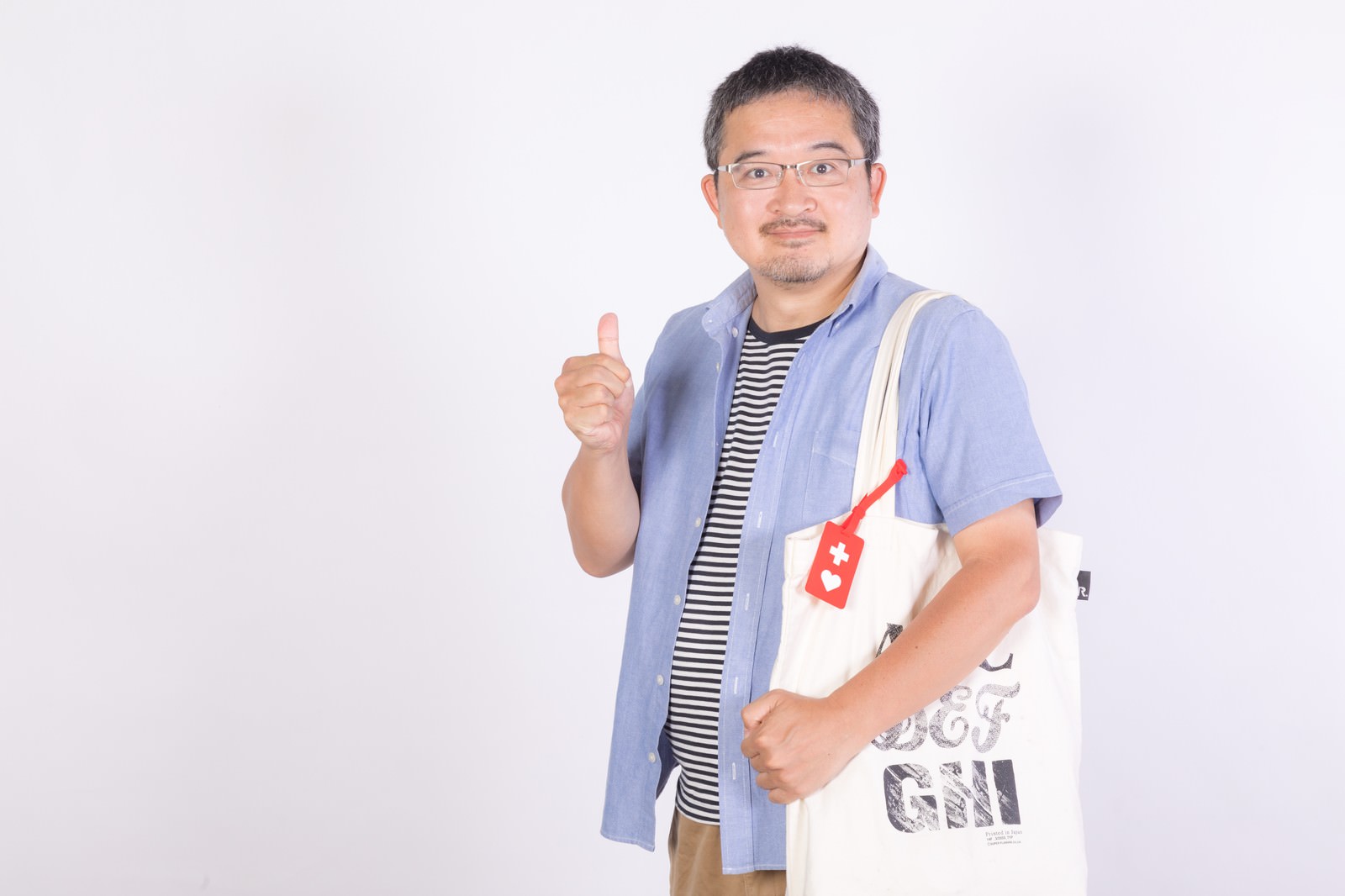 眼鏡をかけた男性が水色シャツを着て親指を立てるいいねポーズ、胸に赤いヘルプマーク［モデル：ナリシゲ］