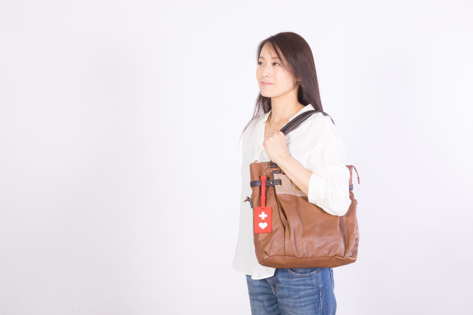 茶色のバッグを肩にかけてヘルプマークをつけた女性が白背景で横向きに立つ［モデル：土本寛子］