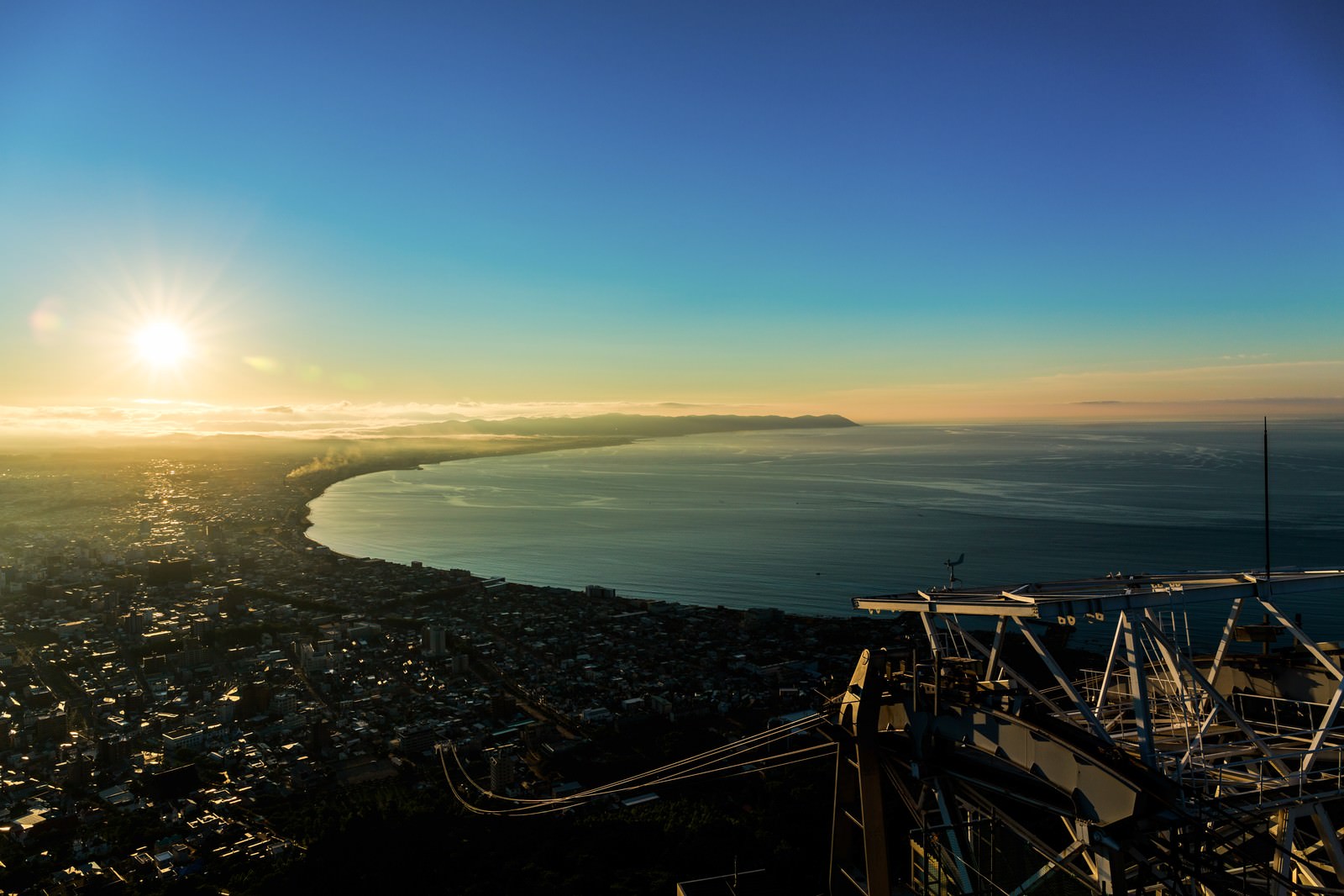 The sunrise over a bay city, as seen from the railing of an observation deck. The sun is rising on the horizon, casting its light over the coastal city below.
