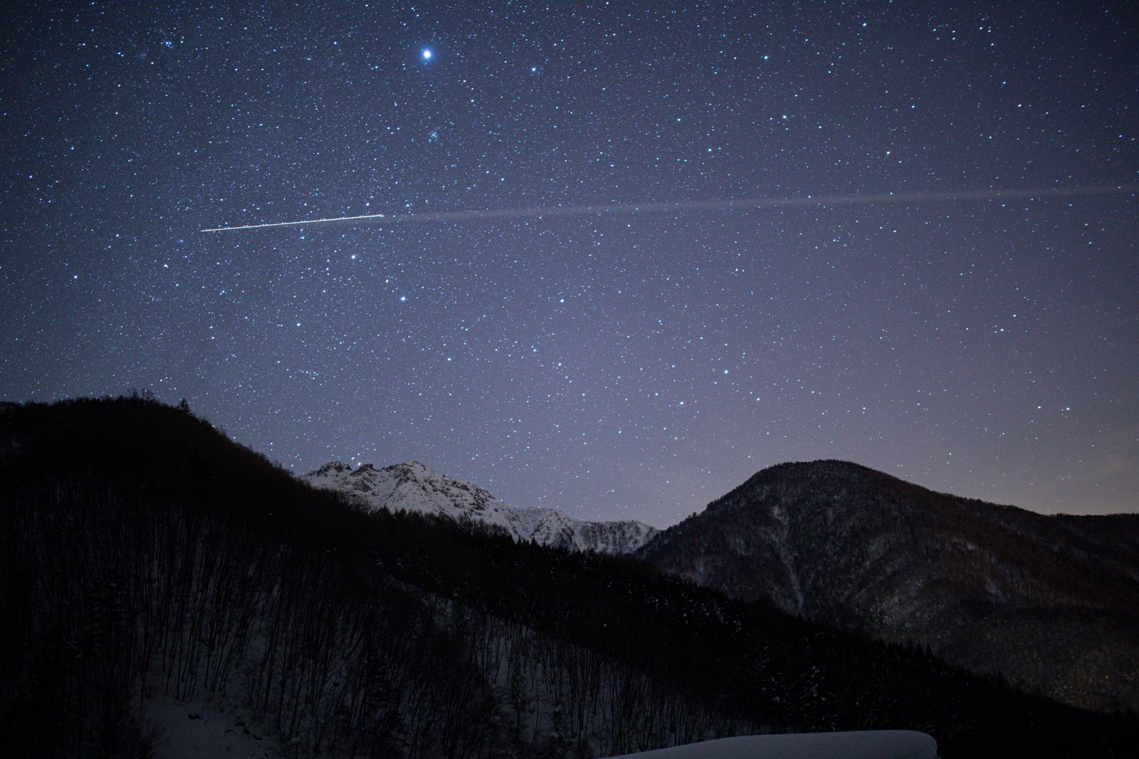 冬の雪山を背景に、満天の星が輝く夜空と流れ落ちる流星