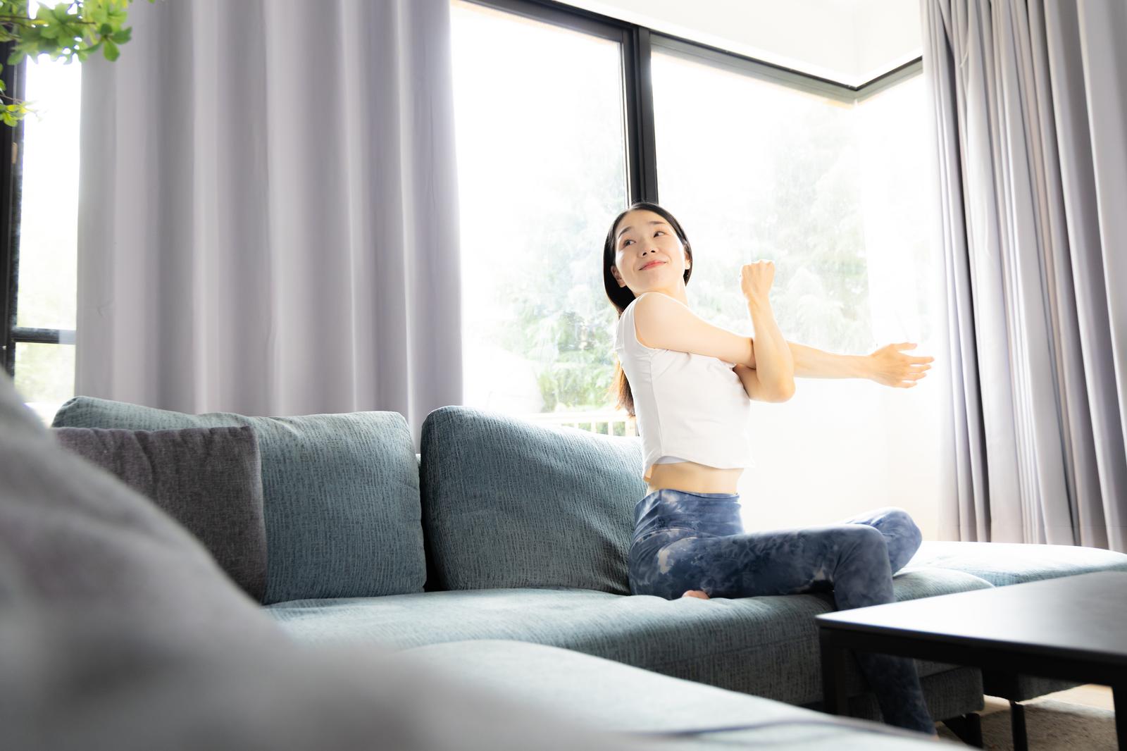 A woman sitting on the sofa in a living room is stretching her arms out wide, with a large window and light purple curtains in the background.