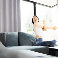 大きな窓のあるリビングでソファーの上でストレッチする女性の写真