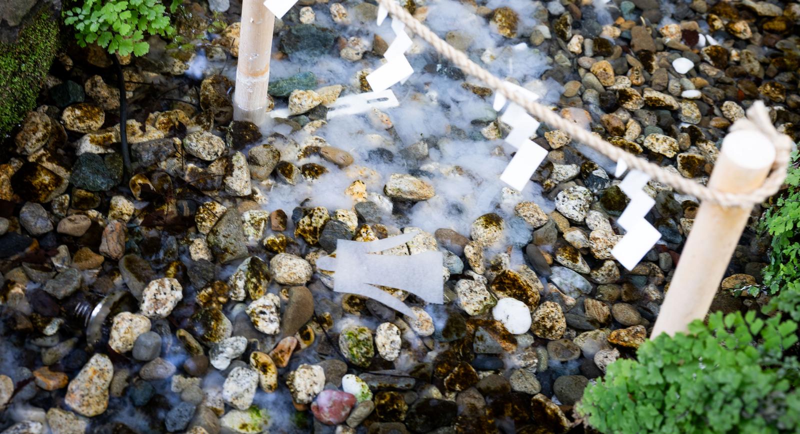 Ningyō nagashi (doll floating) at Hikawa Shrine showing white paper fragments floating in water and scattered shells and pebbles on the riverbed