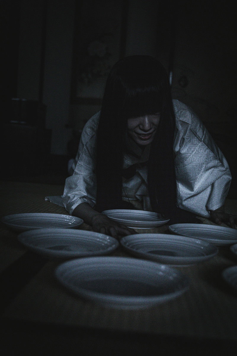 暗い部屋で食器棚の前にうずくまり皿を数える女性［モデル：緋真煉］