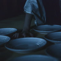 薄暗い和室の畳の上に青白い皿を敷き詰める女性の写真