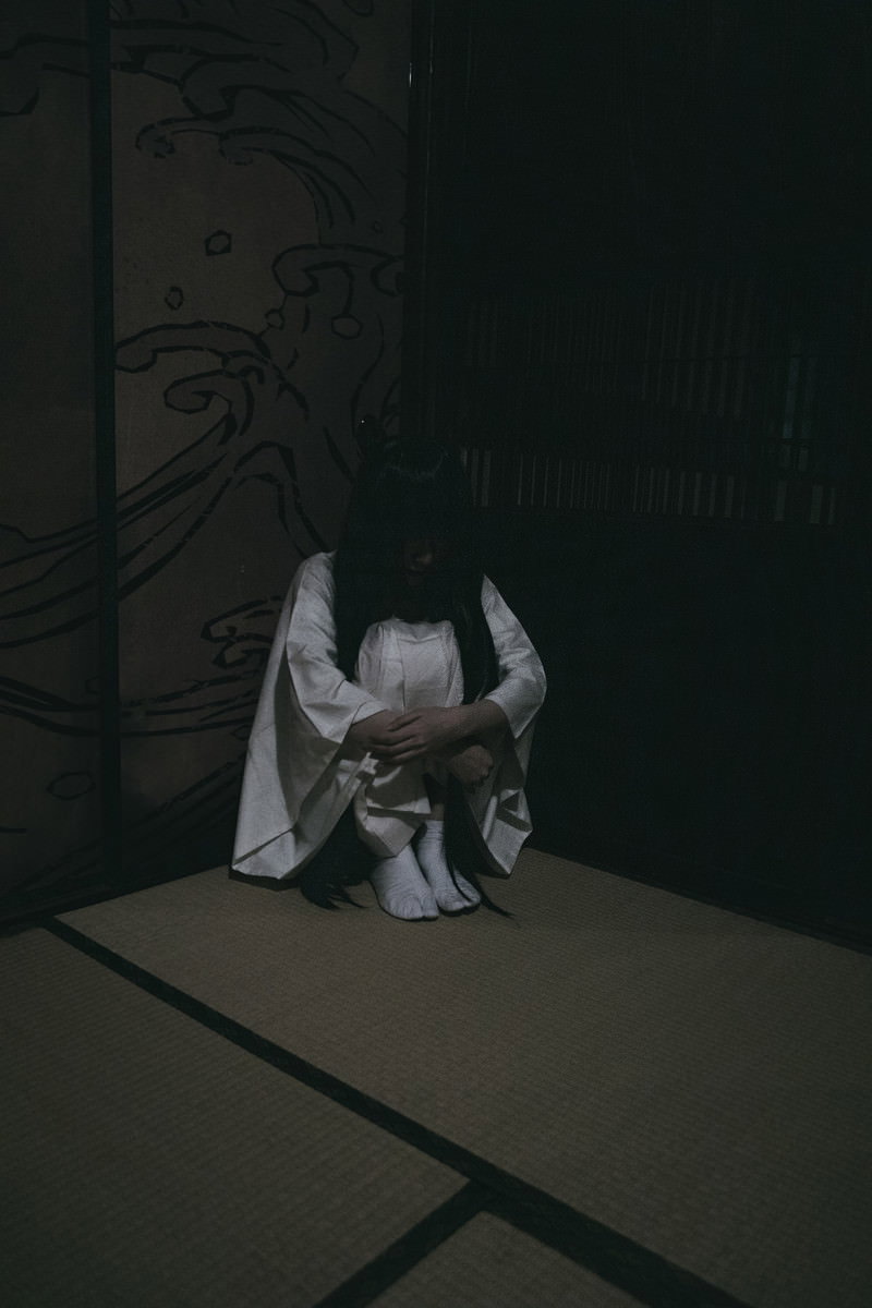 暗い部屋の隅で体育座りをしている人物［モデル：緋真煉］