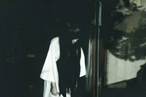 窓際で俯く白い服の女性、暗い室内で窓からの淡い光に照らされる