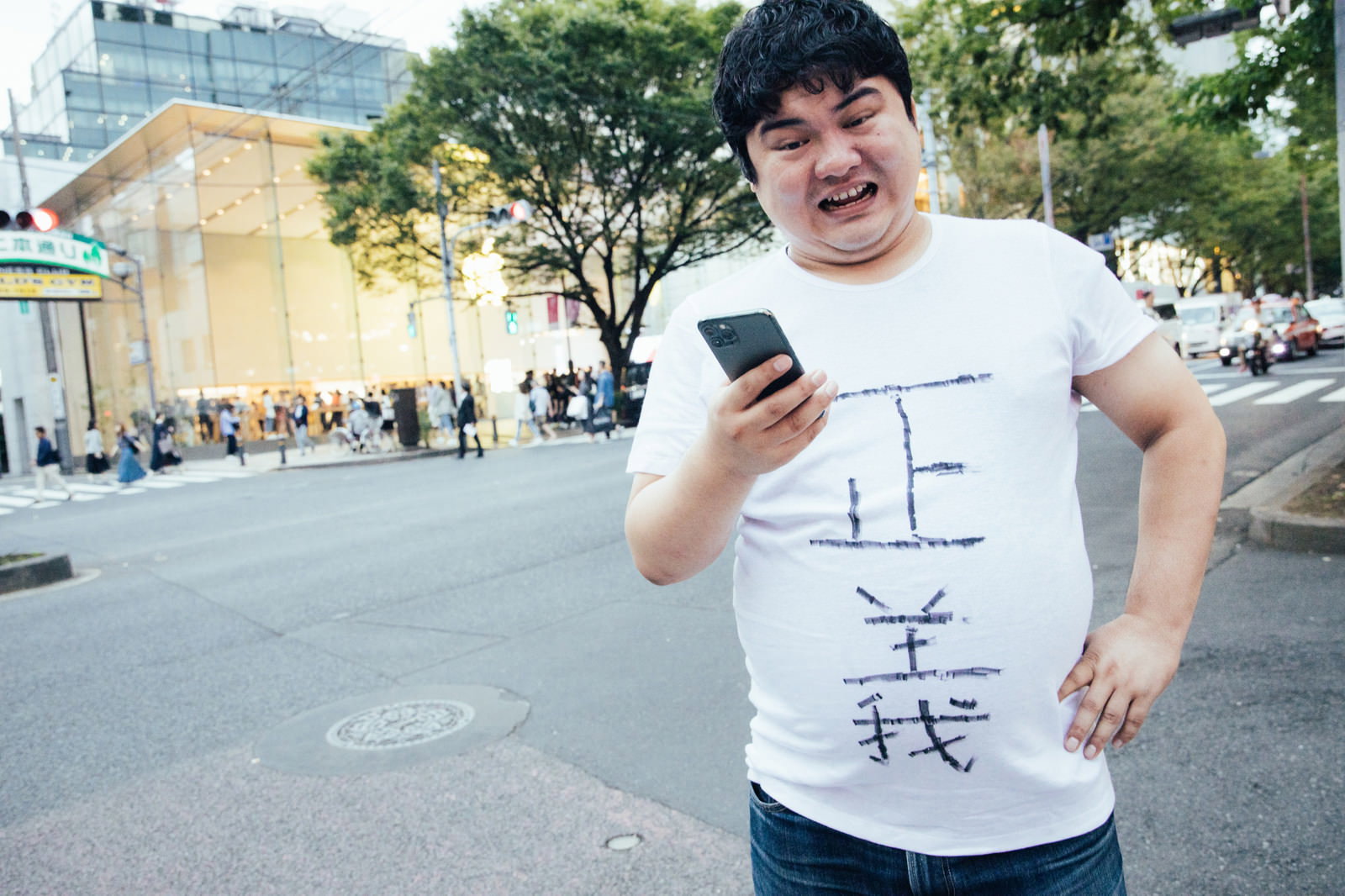 白いTシャツを着た男性が屋外でiPhoneを持ちながら手を広げて説明している様子［モデル：段田隼人］
