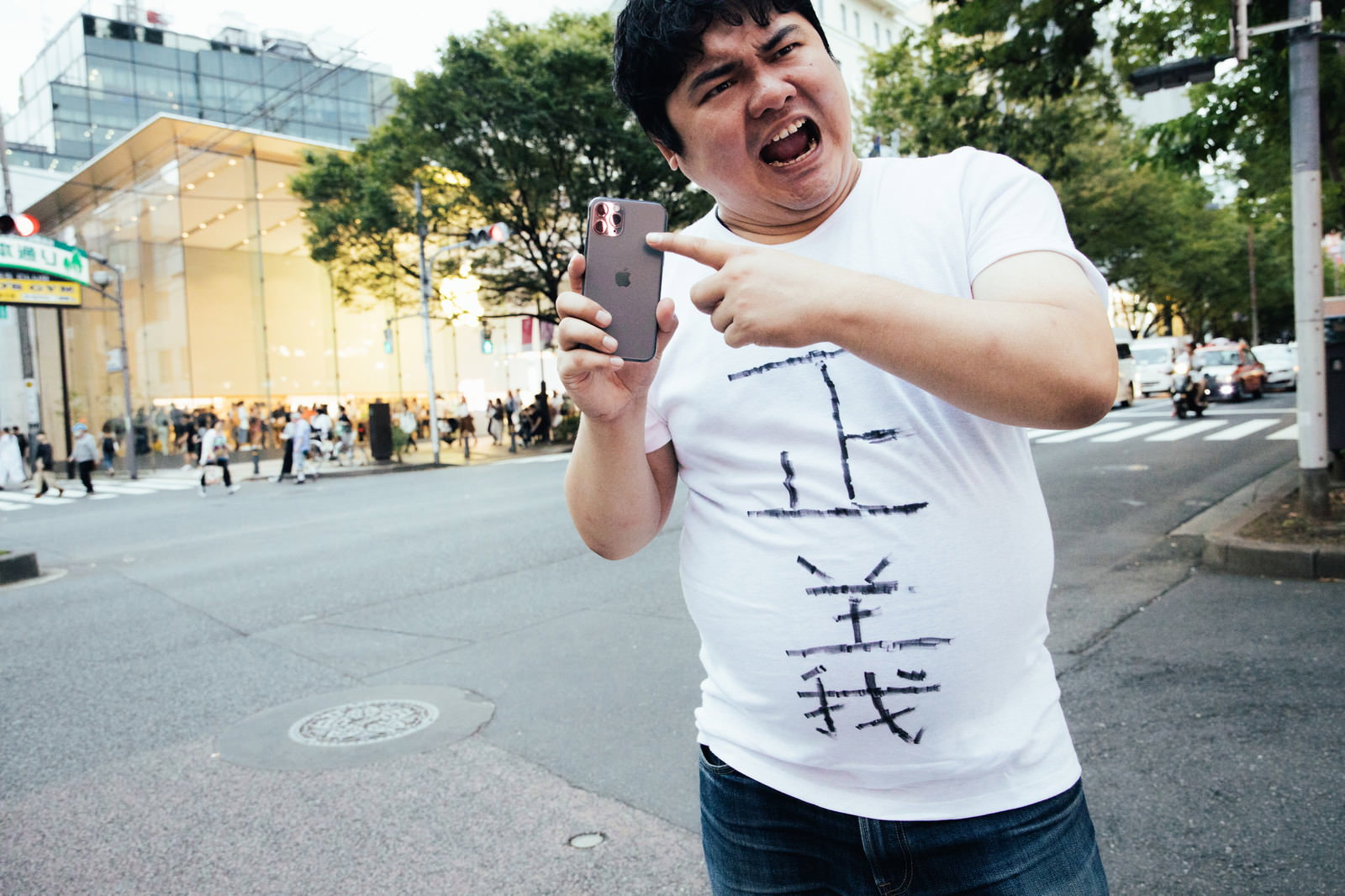 スマートフォンを持ちながら指をさして主張する男性の屋外写真。都市背景［モデル：段田隼人］