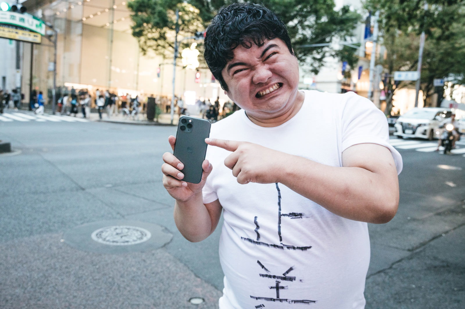 街中で白いTシャツを着た男性が両手でiPhone11を持ち、人差し指を立てながら怒った表情で抗議している［モデル：段田隼人］
