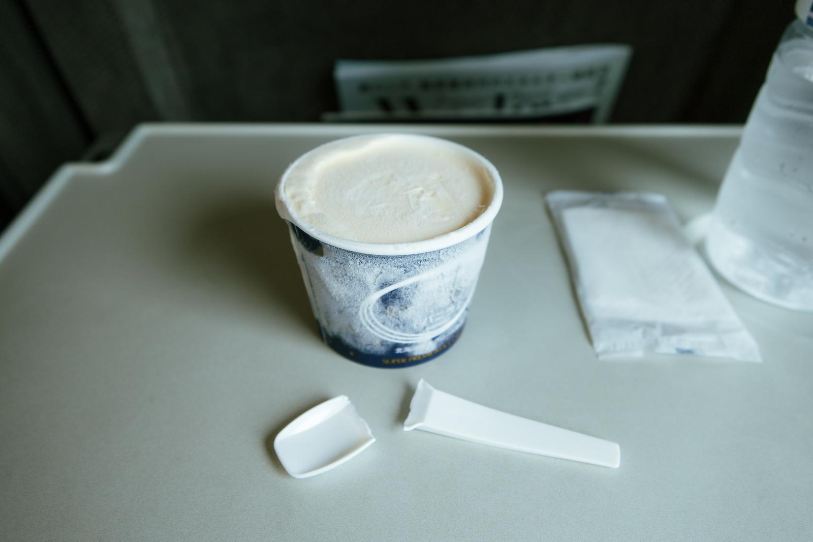飛行機の白いトレーの上に乗った凍ったバニラアイスと折れたプラスチックスプーン
