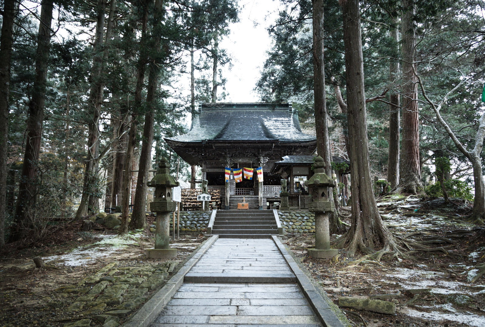 雪の降る中尊寺境内に建つ朱色の弁慶堂と雪化粧した杉並木