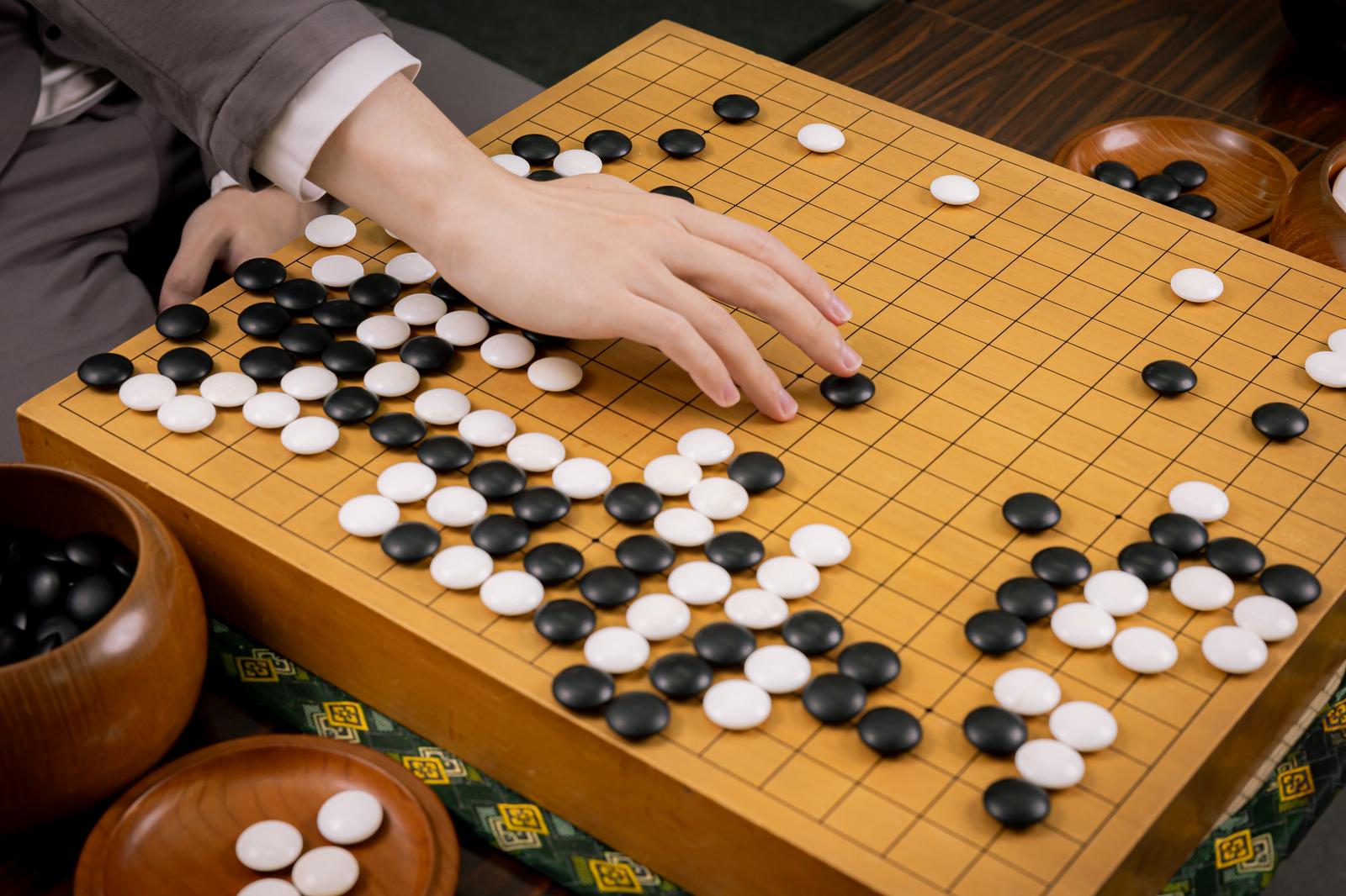 木製の碁盤に白石と黒石が打たれた囲碁の盤面で、棋士の手が白石を置こうとしている