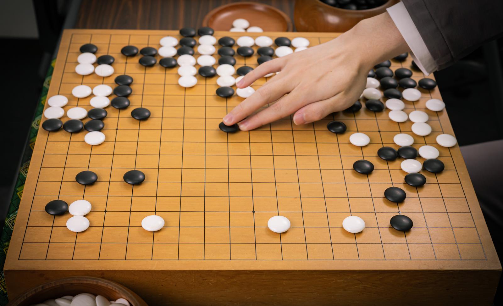 木製の碁盤に黒石と白石が並ぶ囲碁の対局で、プレイヤーが石を置こうとしている［モデル：囲碁］