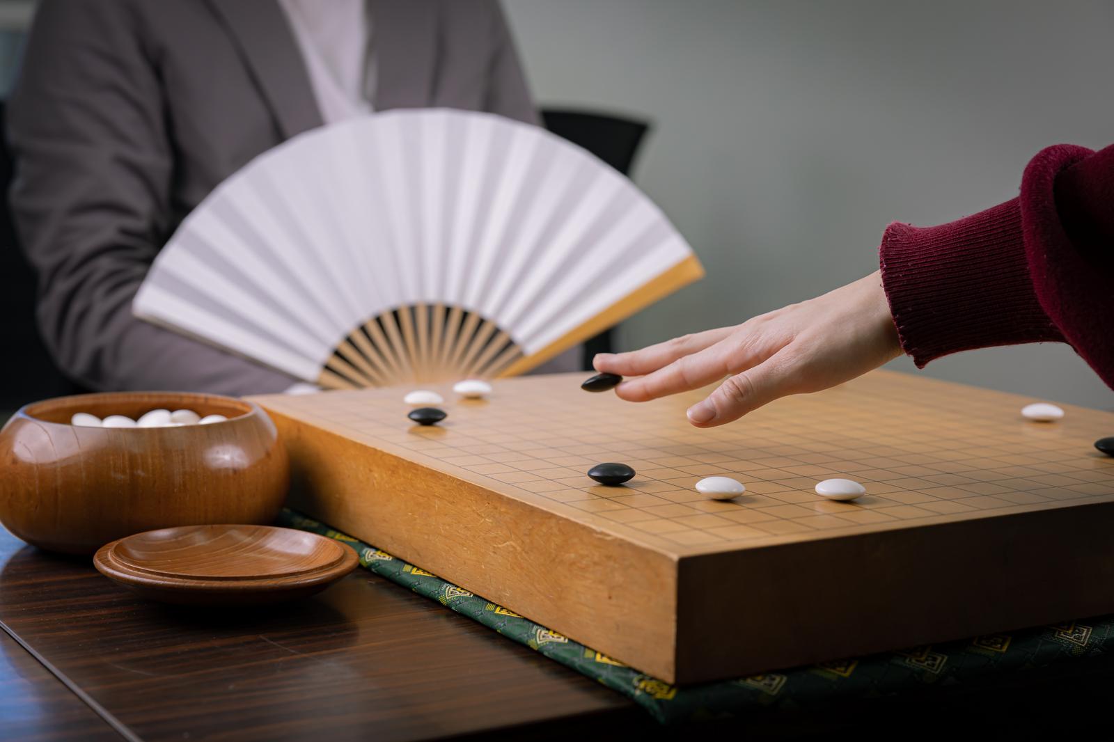 碁盤に向かう2人の囲碁棋士。片方が扇子を広げて持ち、碁石を配置している